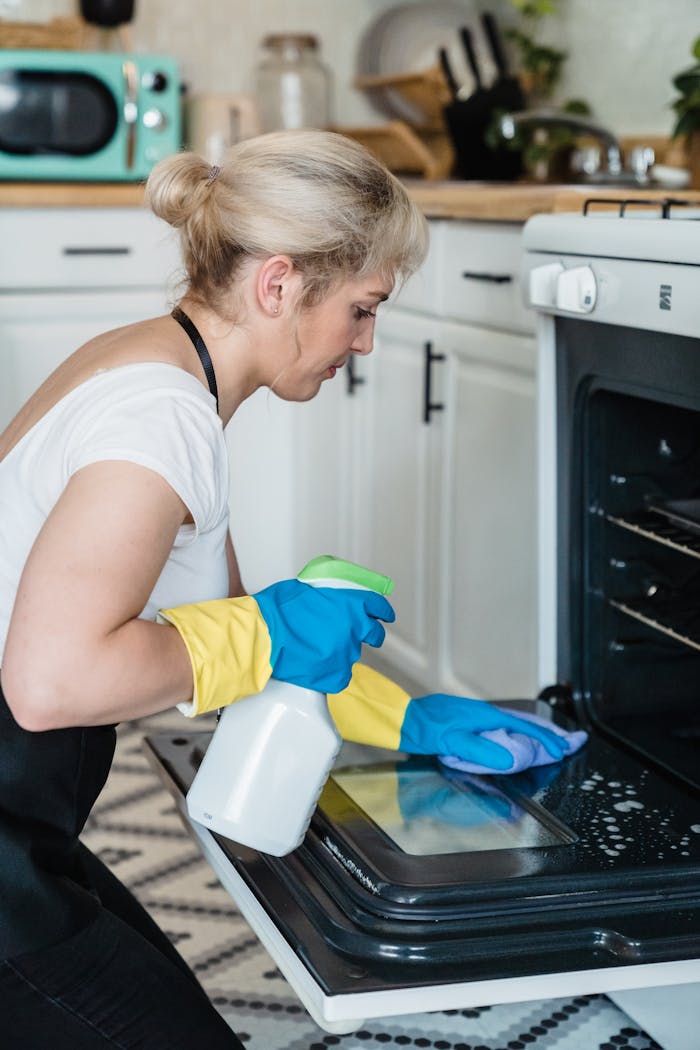 about-us A woman uses a spray bottle and cloth to clean an oven in a modern kitchen setting.